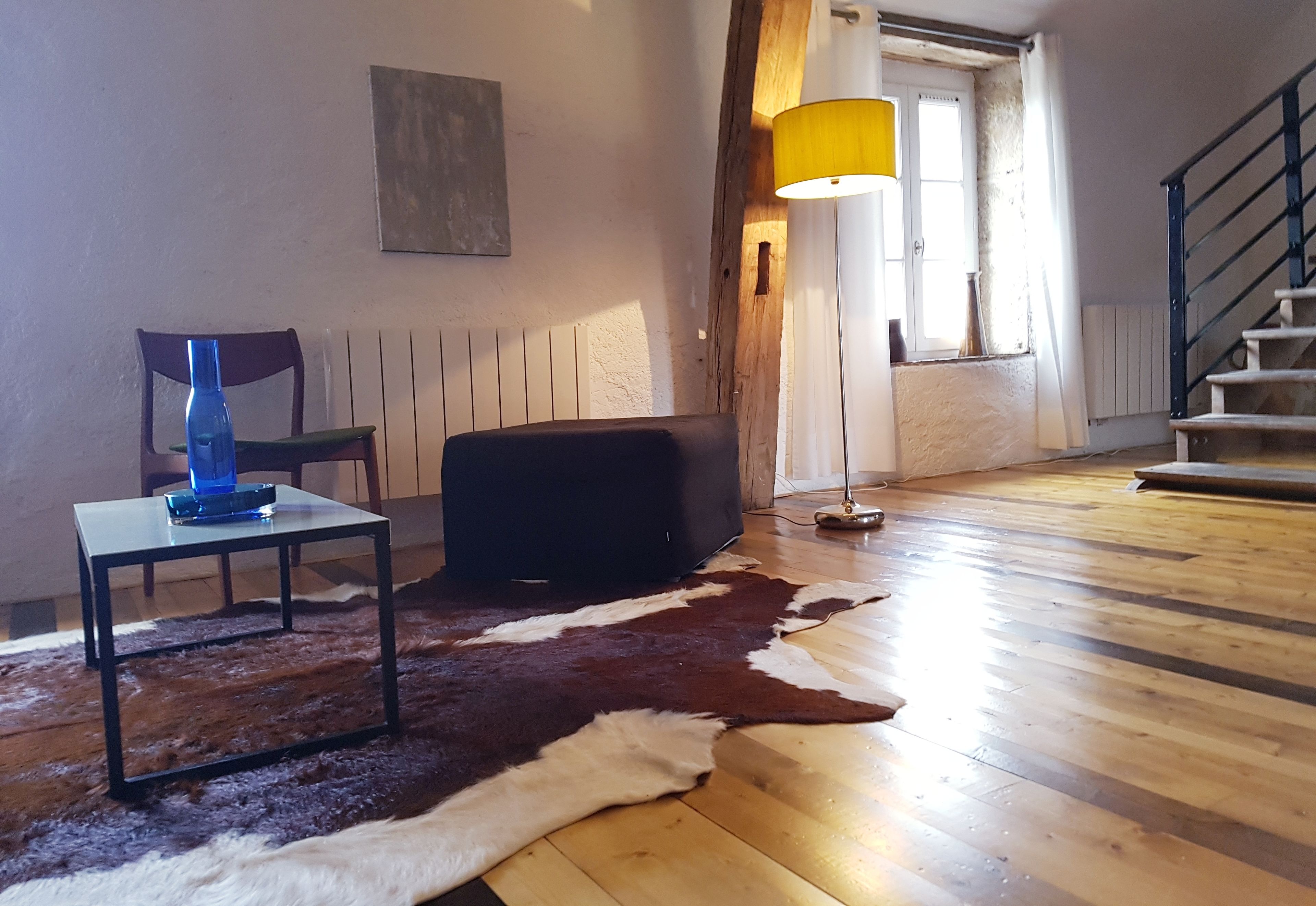 A room with a wooden floor, a black ottoman, a blue bottle on a small table, a cowhide rug, a lamp with a yellow shade, a staircase, and a radiator.