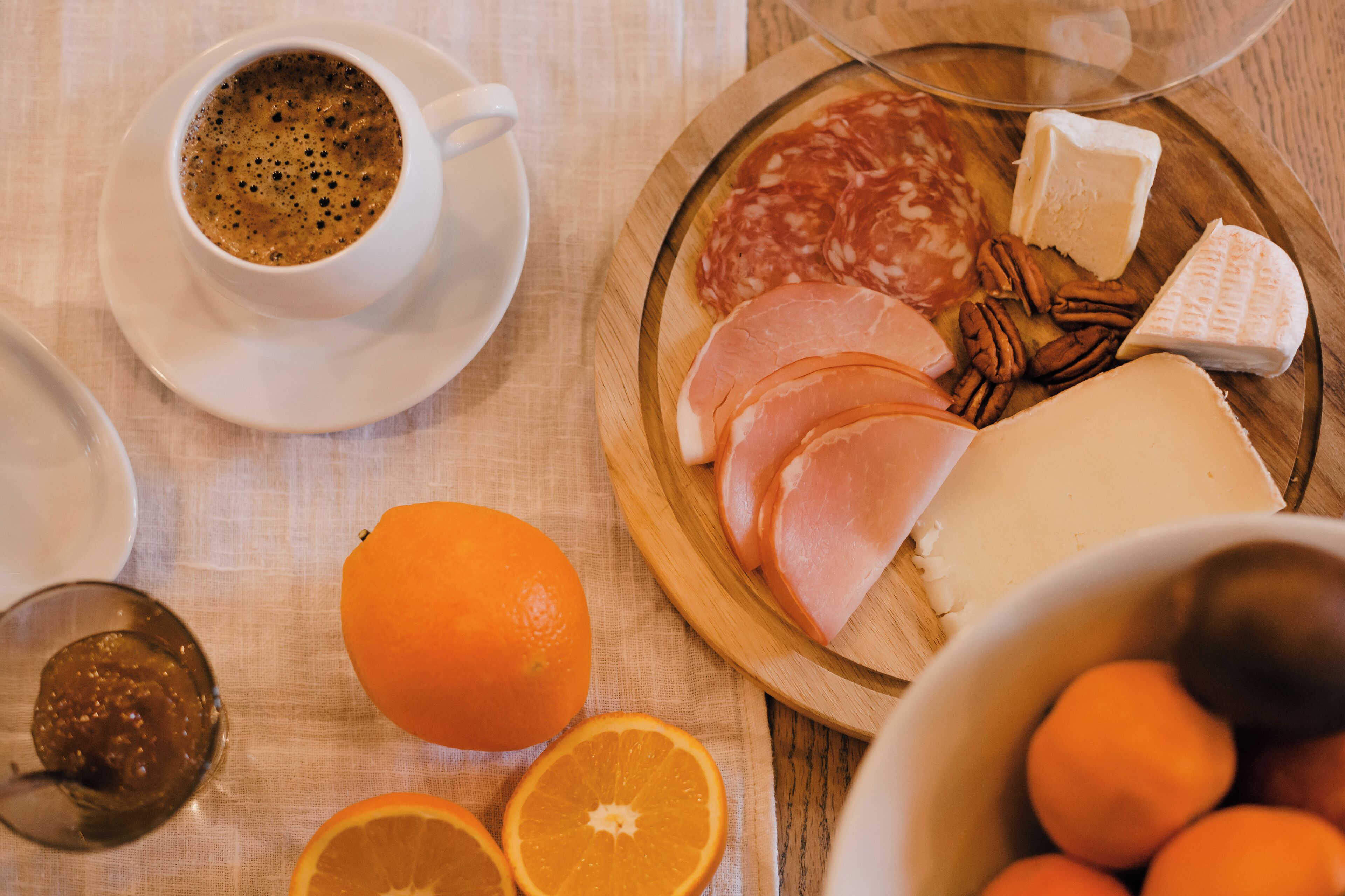 A breakfast setting with a cup of coffee, a plate of ham, cheese, and oranges on a wooden board.