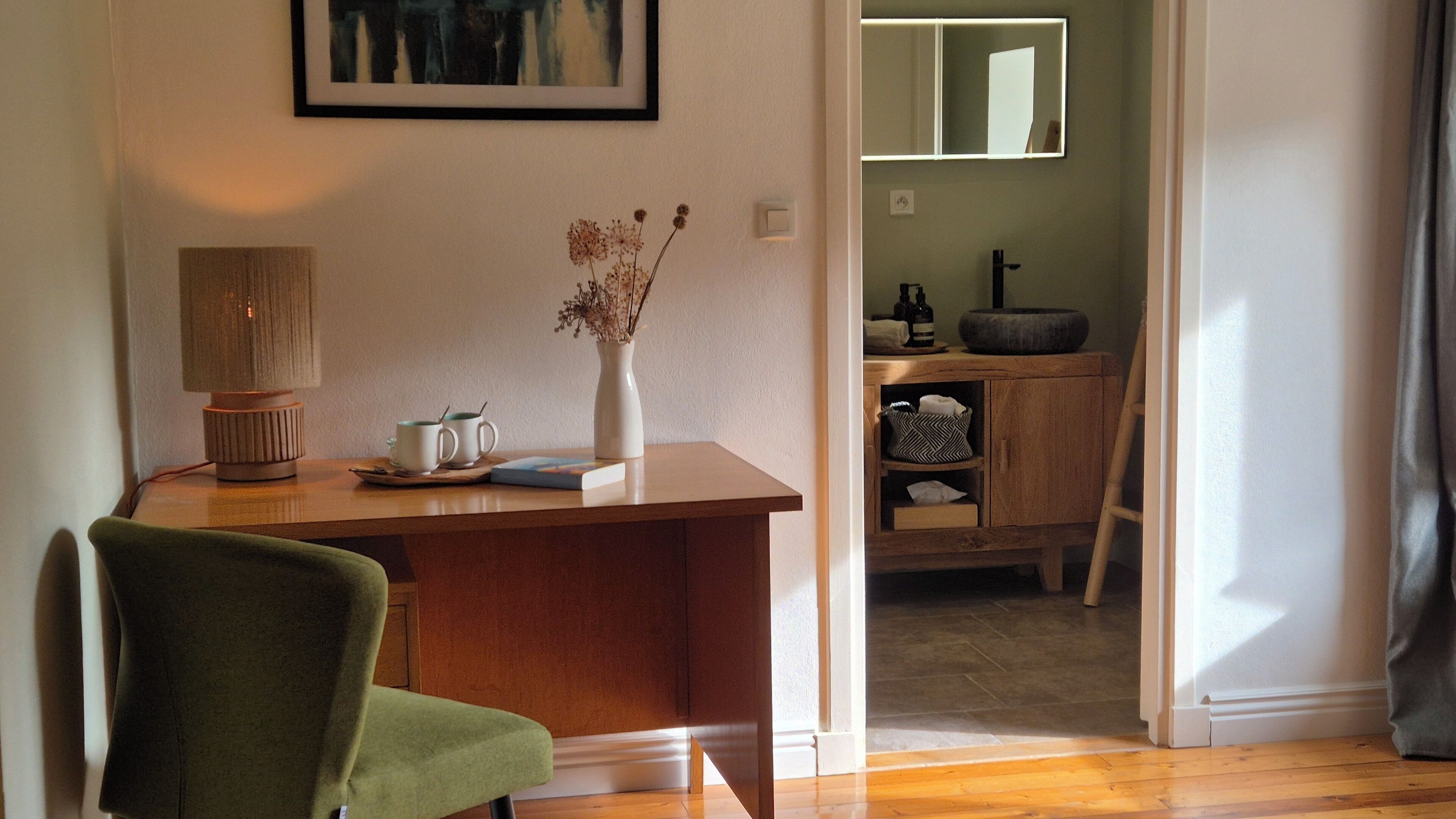 A room with a wooden desk, a chair, a lamp, a vase with dried flowers, and a mirror on the wall.