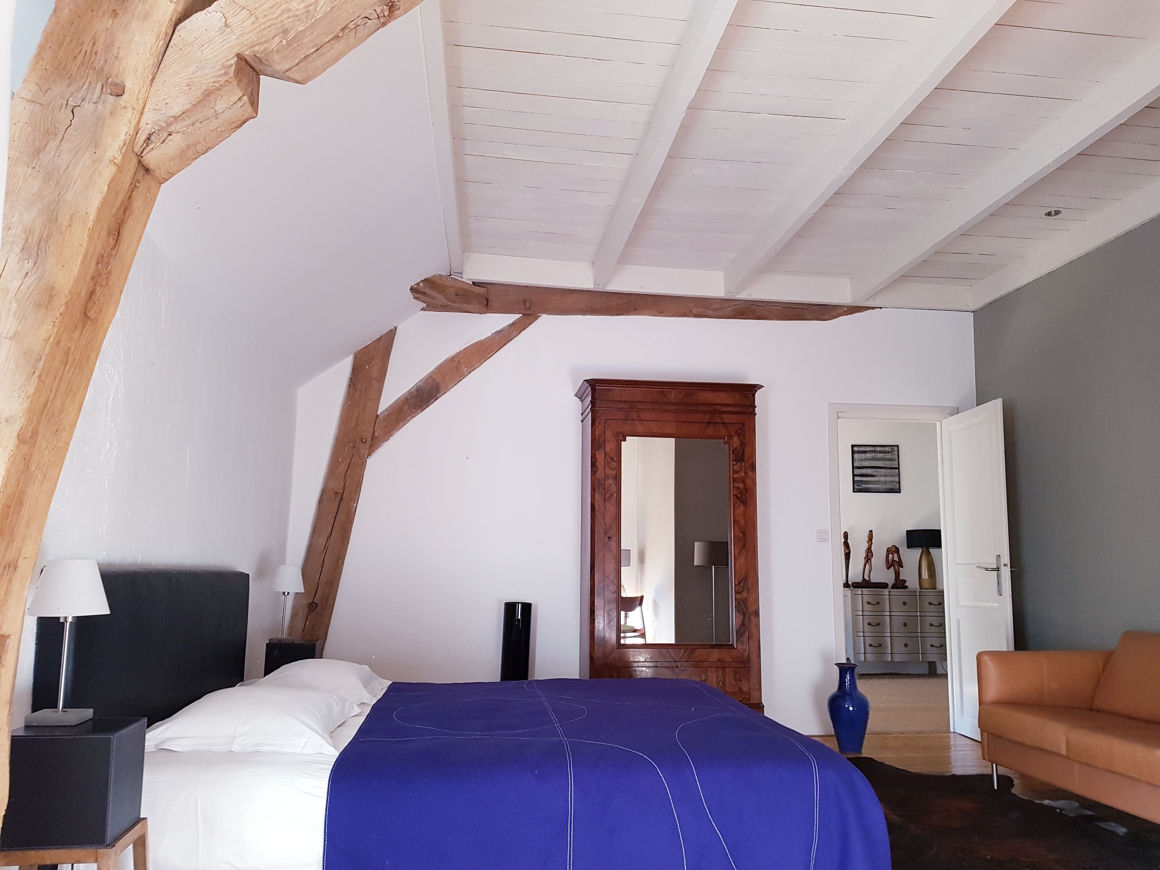 Een slaapkamer met een schuine plafond, een bed met een blauwe spreideken, een houten hoofdbord, een nachtkastje met een lamp, een dressoir en een deur naar een andere kamer.