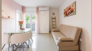 Un salon moderne avec un canapé beige, une table à manger en verre et deux chaises, une petite table d’appoint sur laquelle est posé un vase de fleurs, une étagère murale et un tableau.