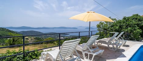 A terrace with white plastic lounge chairs and a yellow umbrella overlooking a body of water and distant mountains.