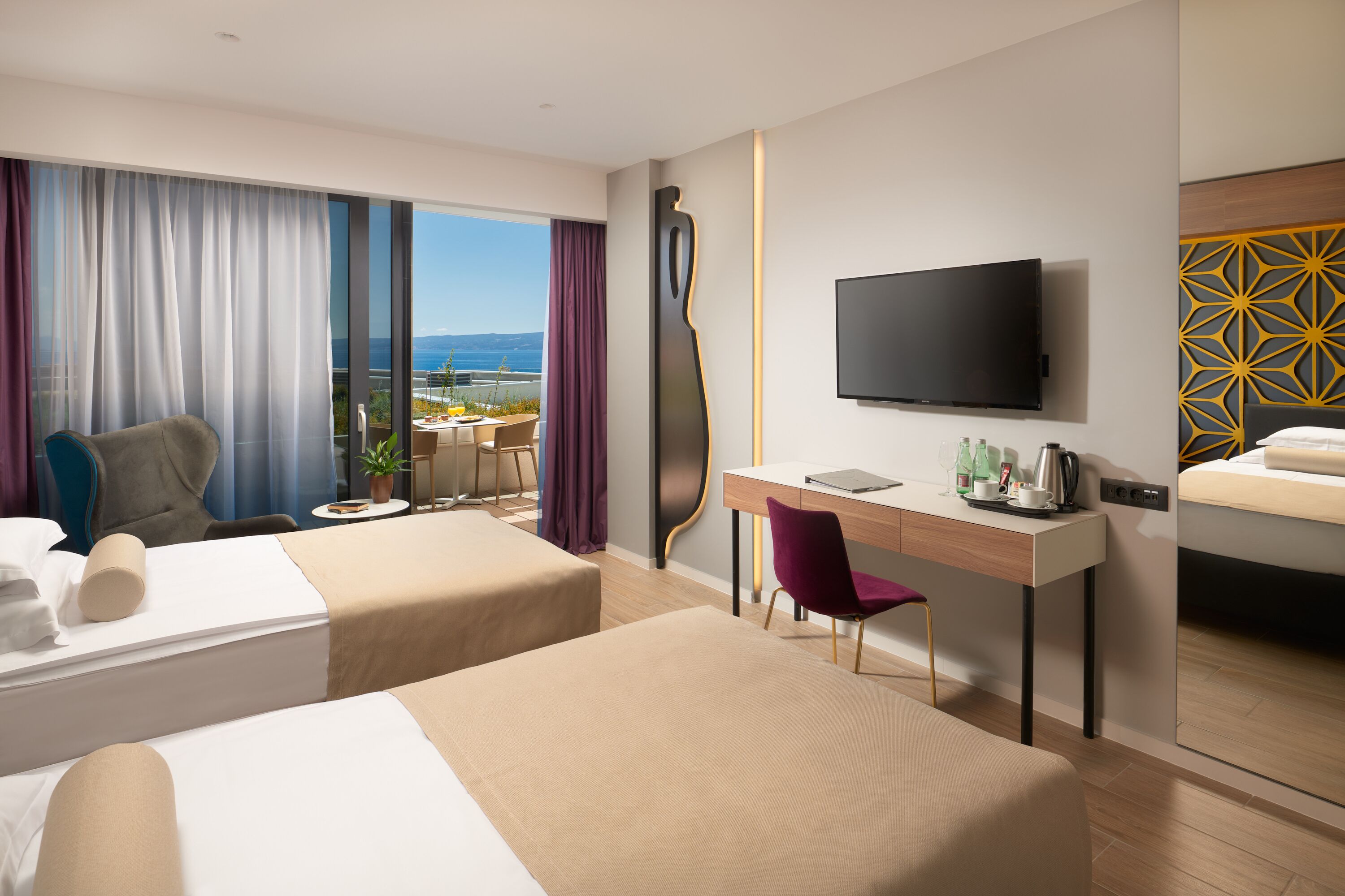 A hotel room with two beds, a desk, a chair, a TV, and a view of the sea.