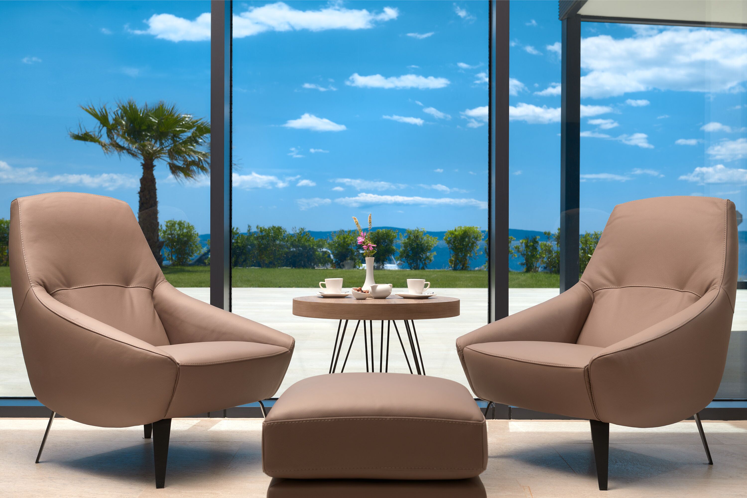Twee beige fauteuils gericht op een ronde tafel met een vaas en kopjes, voor een groot raam met uitzicht op palmbomen en een heldere lucht.