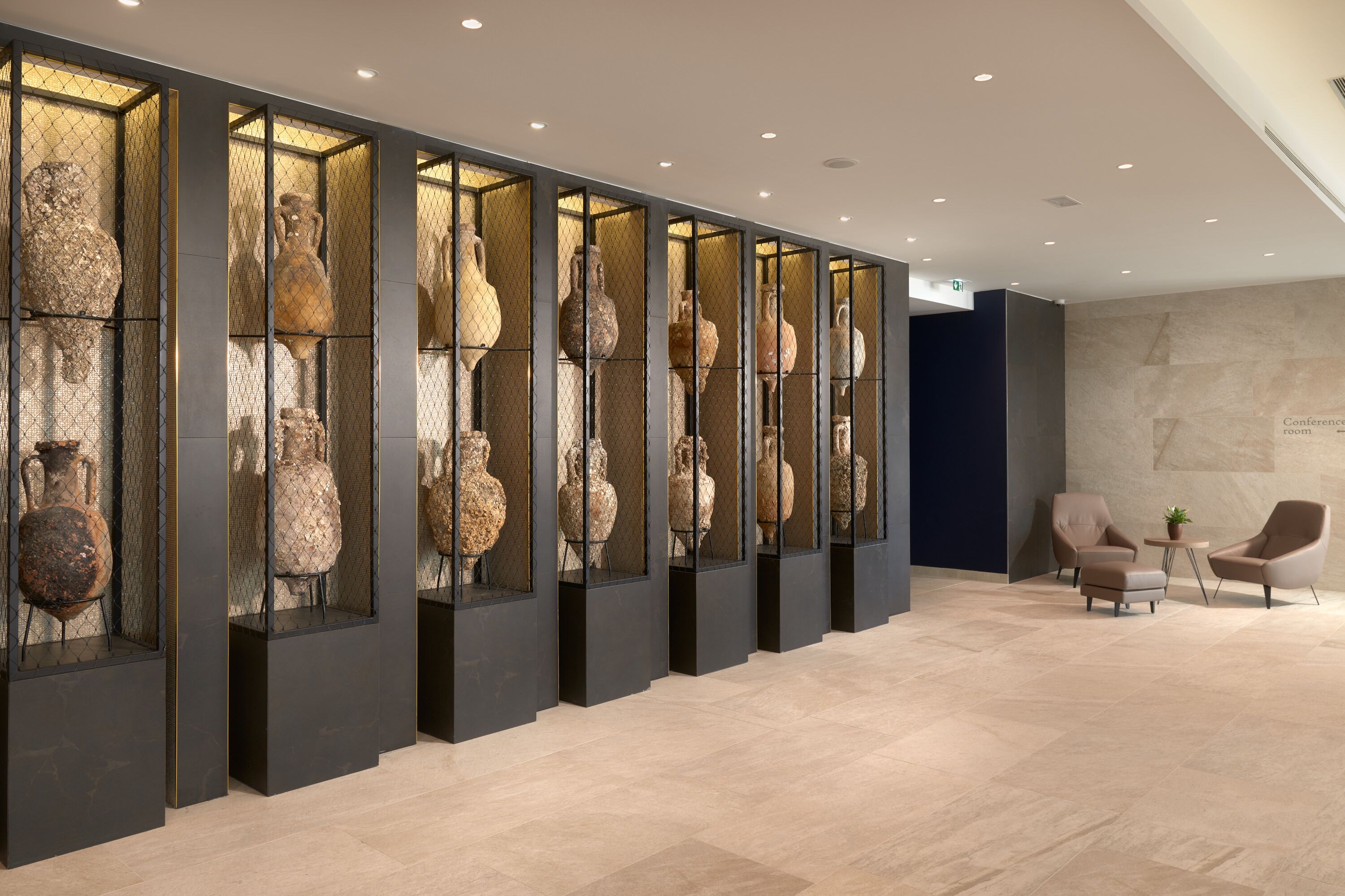 Een moderne lobby met een rij vitrines vol oude potten, een zithoek met twee fauteuils en een bijzettafeltje, en een marmeren vloer.