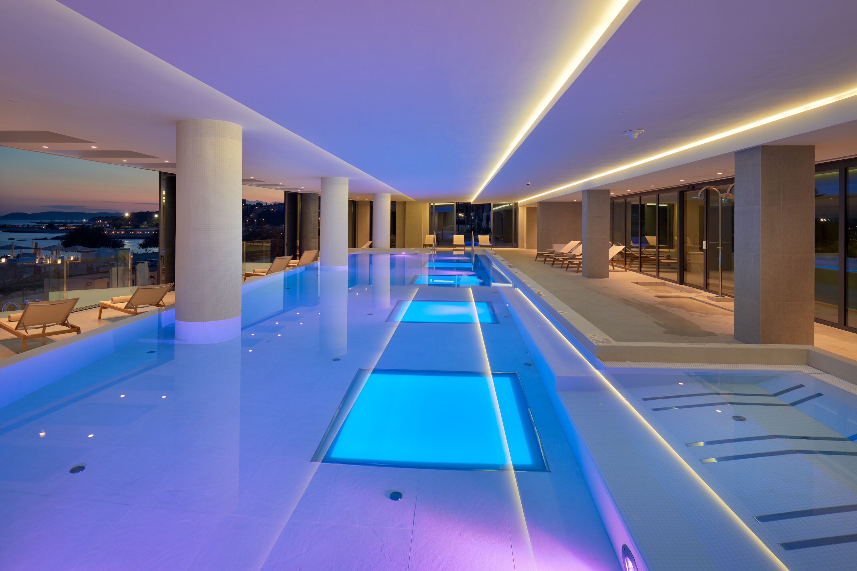 A modern indoor swimming pool with blue lighting, lounge chairs, and a view of the city at dusk.