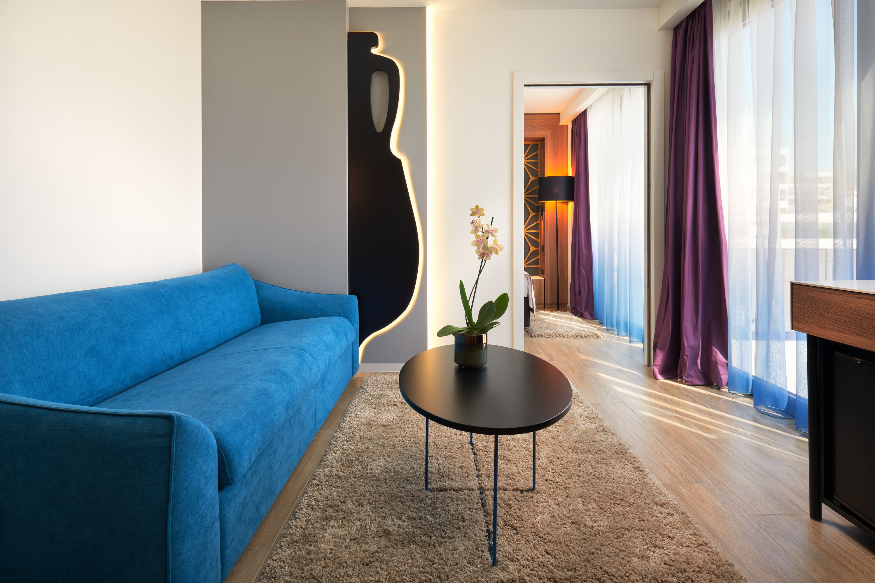 Una sala de estar moderna con un sofá azul, una mesa de centro redonda negra con una planta, y vistas a un pasillo con cortinas moradas.