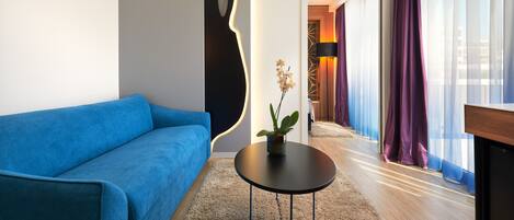 Una sala de estar moderna con un sofá azul, una mesa de centro redonda negra con una planta, y vistas a un pasillo con cortinas moradas.