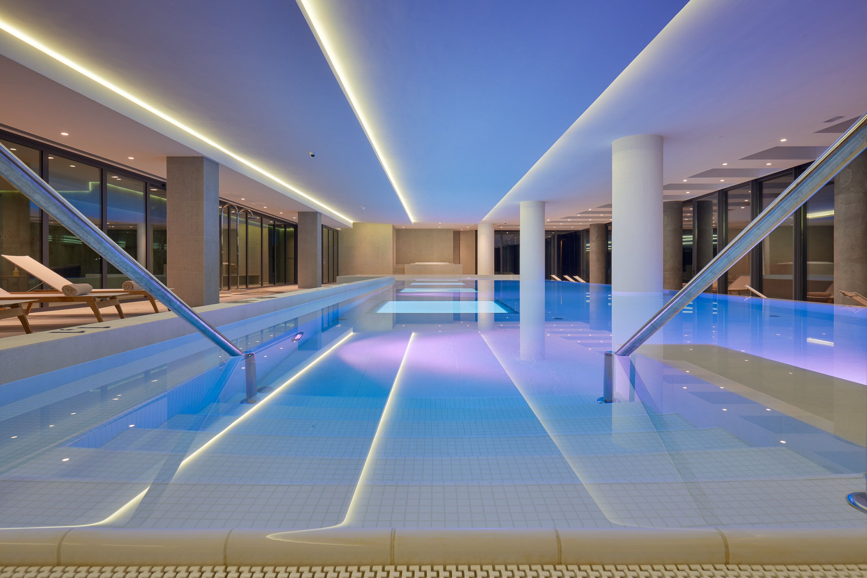 Una piscina interior moderna con agua cristalina, rodeada de paredes de vidrio e iluminada con luces subacuáticas.
