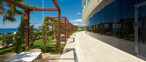 A modern outdoor patio with a long table and chairs, a wooden pergola with greenery, and a glass building with a view of the ocean.
