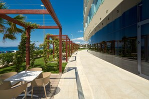 A modern outdoor patio with a long table and chairs, a wooden pergola with greenery, and a glass building with a view of the ocean.