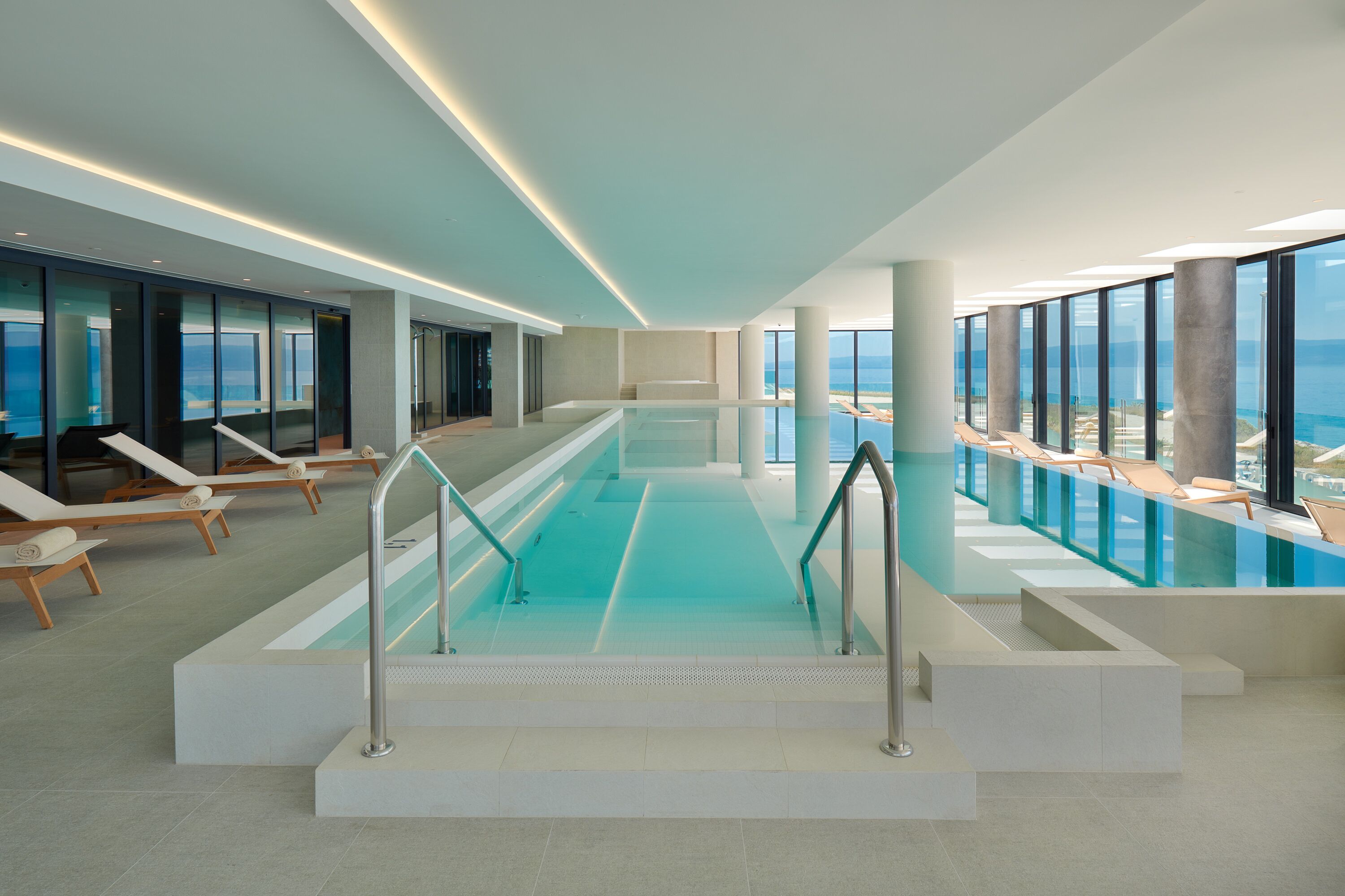 An indoor swimming pool with clear glass walls, lounge chairs, and a view of the ocean.