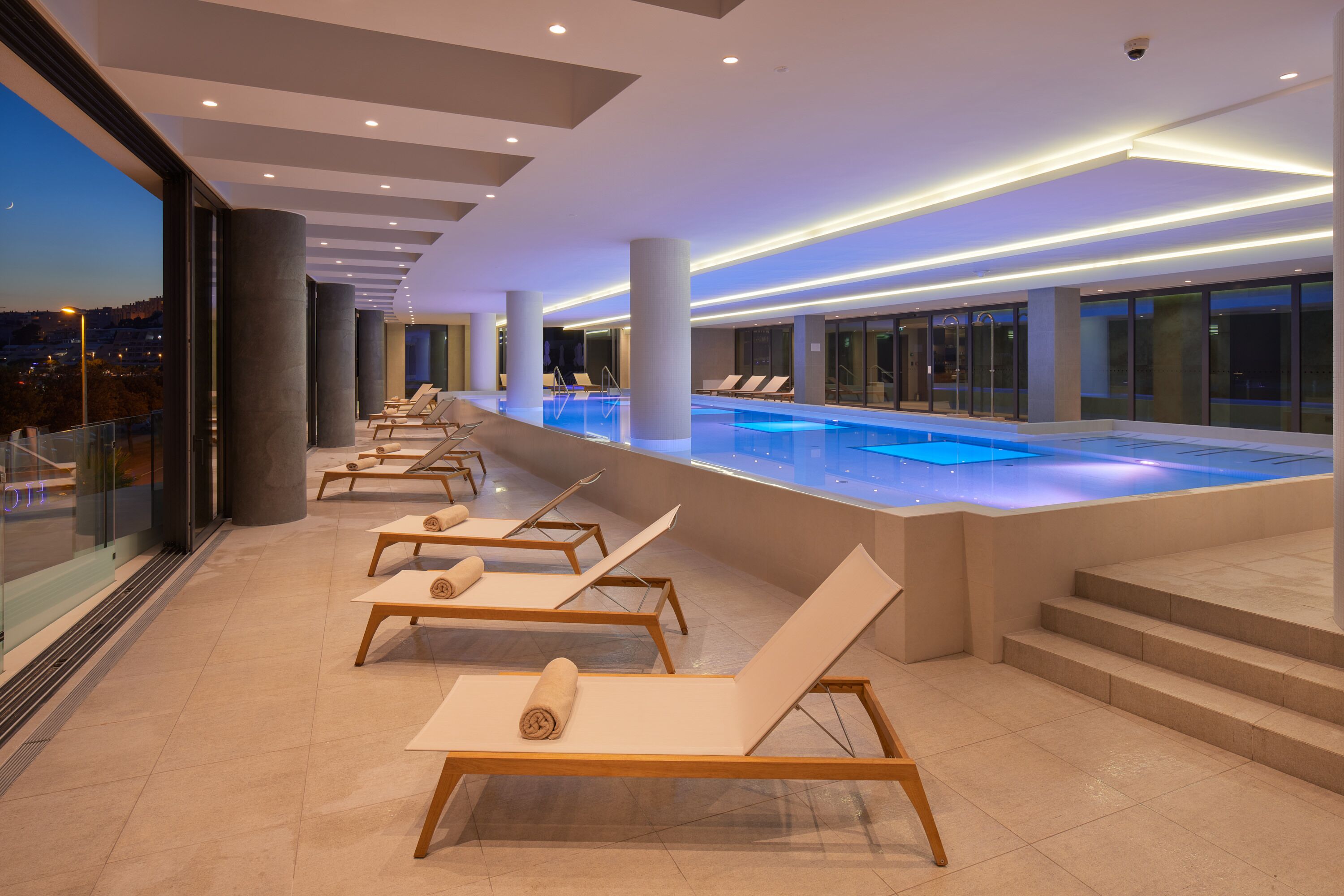 A modern indoor pool area with lounge chairs and a view of the cityscape.