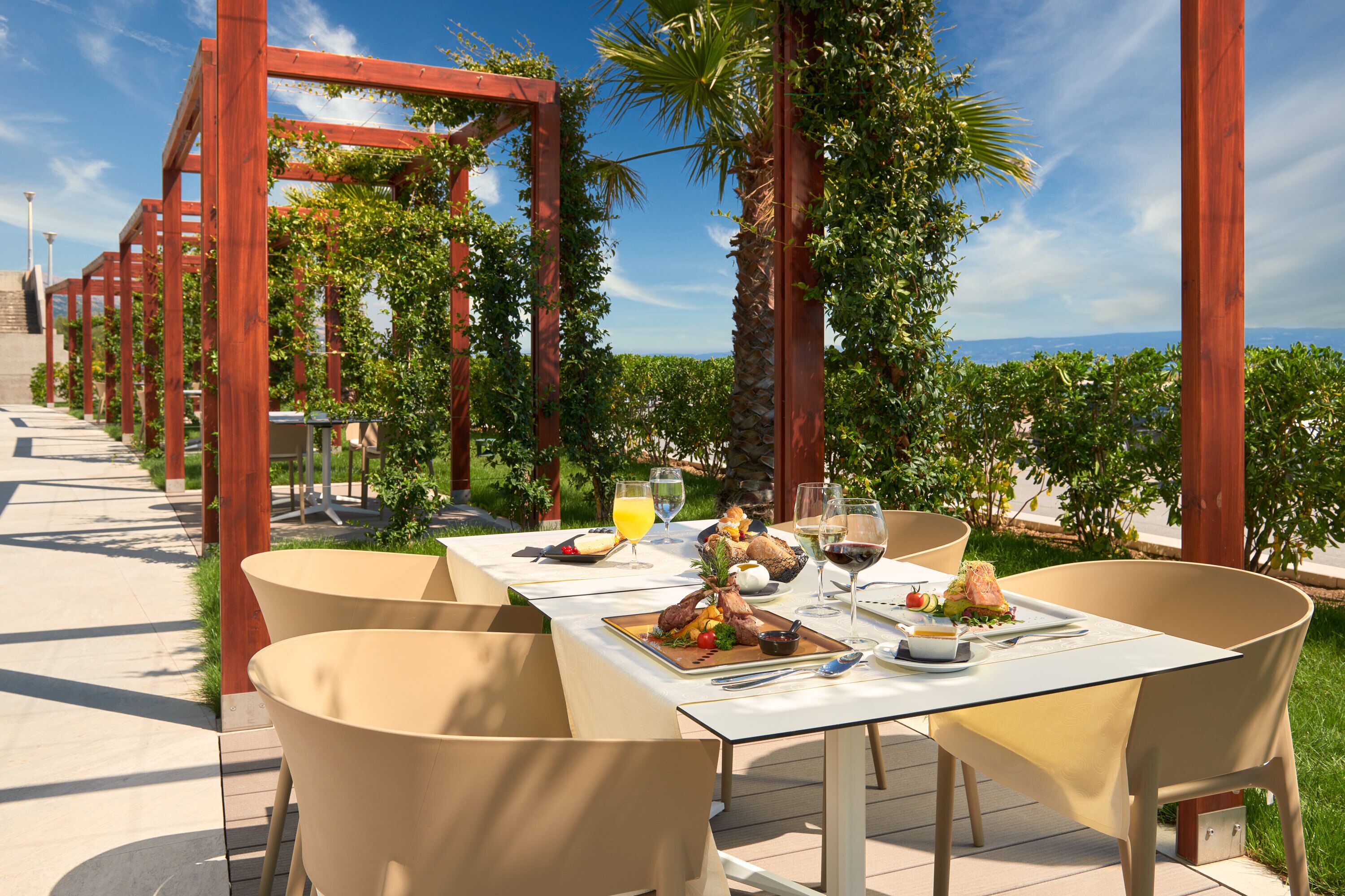 An outdoor dining area with a table set for two, surrounded by chairs, under a wooden pergola with climbing plants.