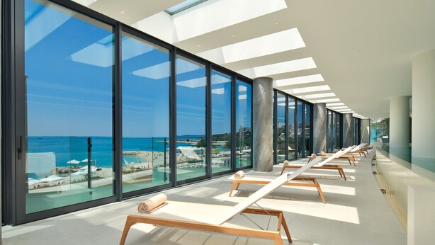 A modern hotel interior with large glass windows offering ocean views, a sunlit corridor with lounge chairs, and a minimalist architectural design.