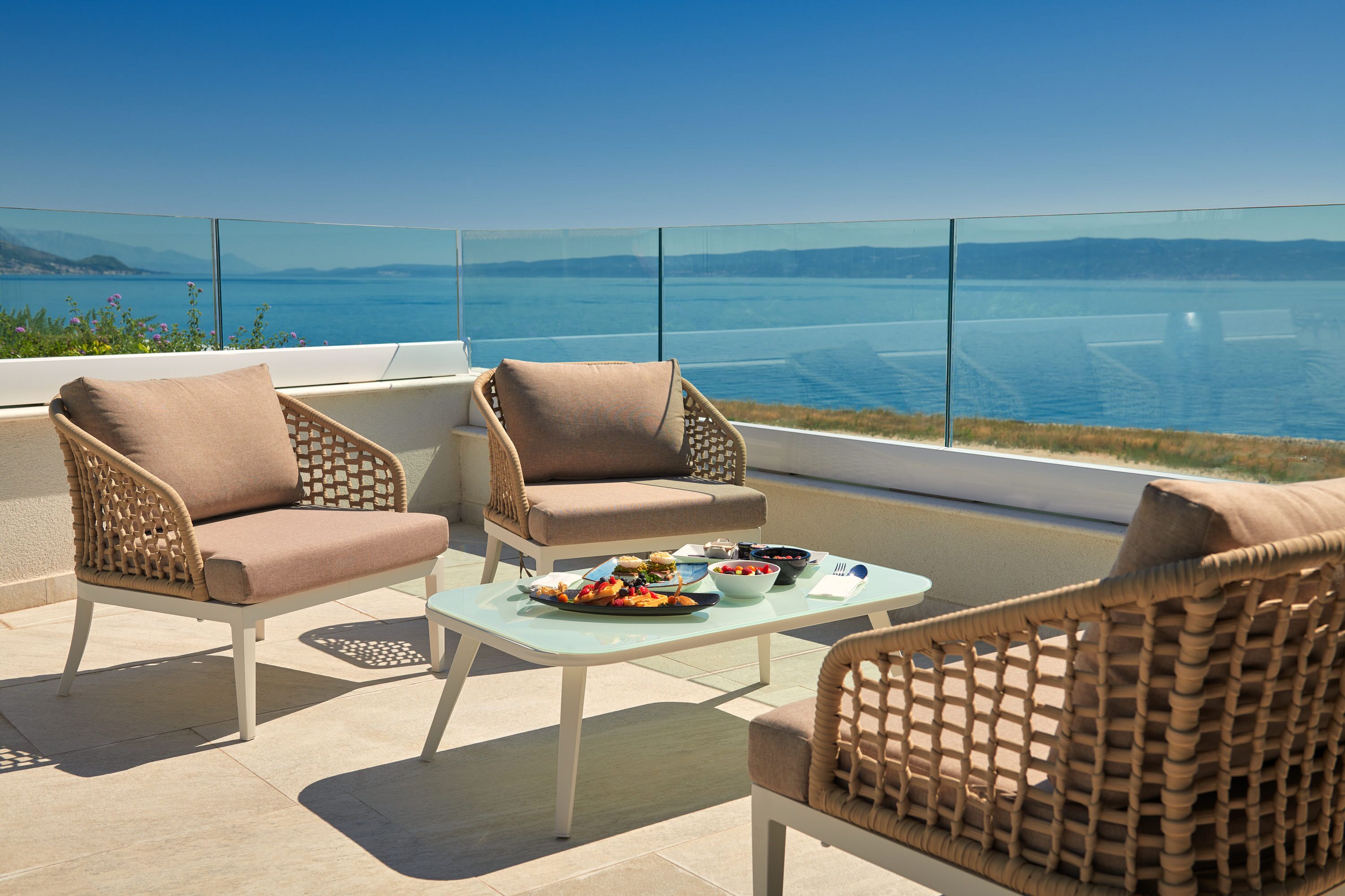 Terraço com cadeiras de vime e uma mesa de vidro, com vista para um corpo de água e montanhas ao longe.