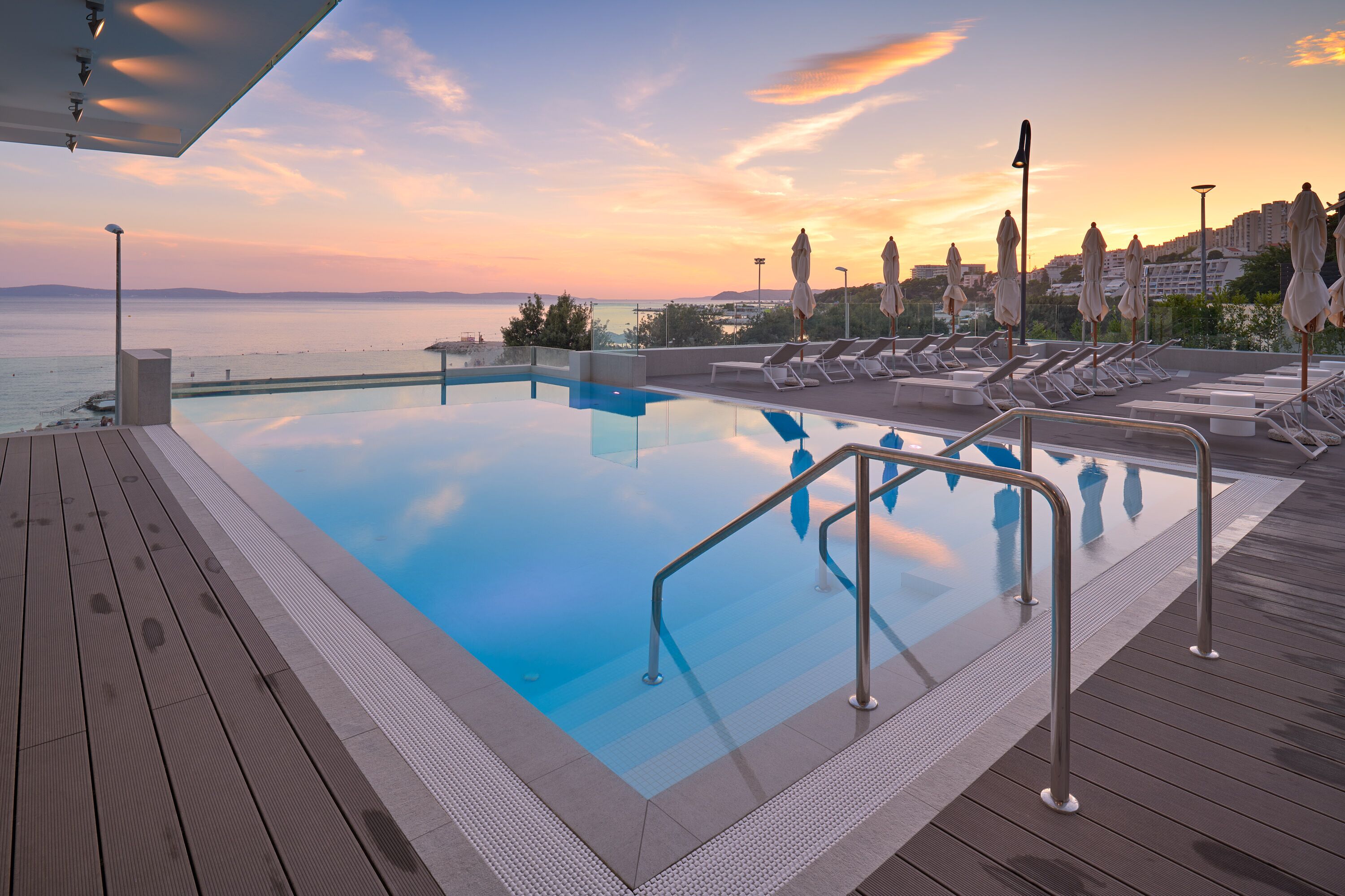 Ein Rooftop-Endlospool mit Liegestühlen und Sonnenschirmen, mit Blick auf eine Stadtlandschaft und das Meer bei Sonnenuntergang.