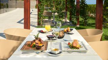 Une table à manger installée à l’extérieur, où sont servis divers plats, notamment des fruits de mer et des légumes, dans un jardin agrémenté de chaises et offrant une vue sur le paysage.