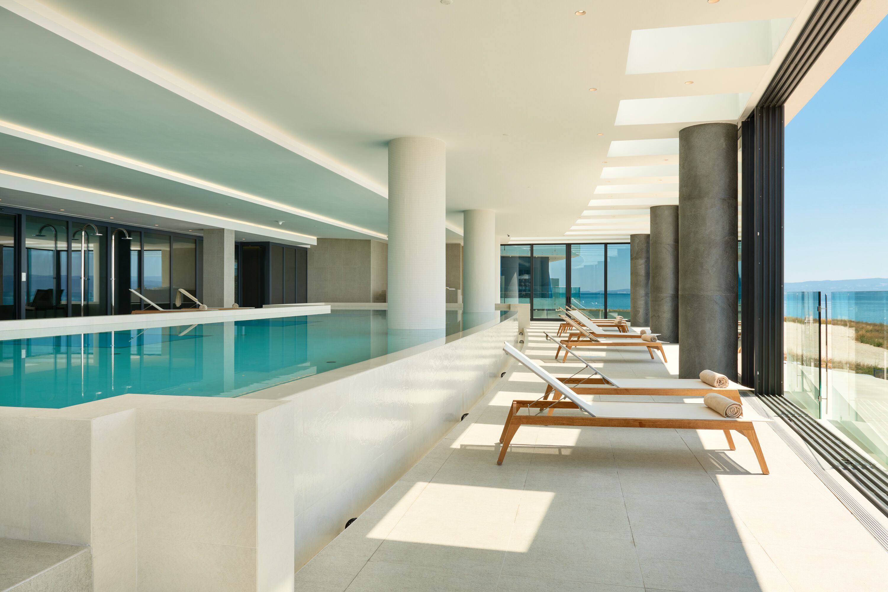 A modern indoor swimming pool with sunlit lounge chairs and large windows overlooking the sea.