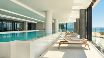 A modern indoor swimming pool with sunlit lounge chairs and large windows overlooking the sea.