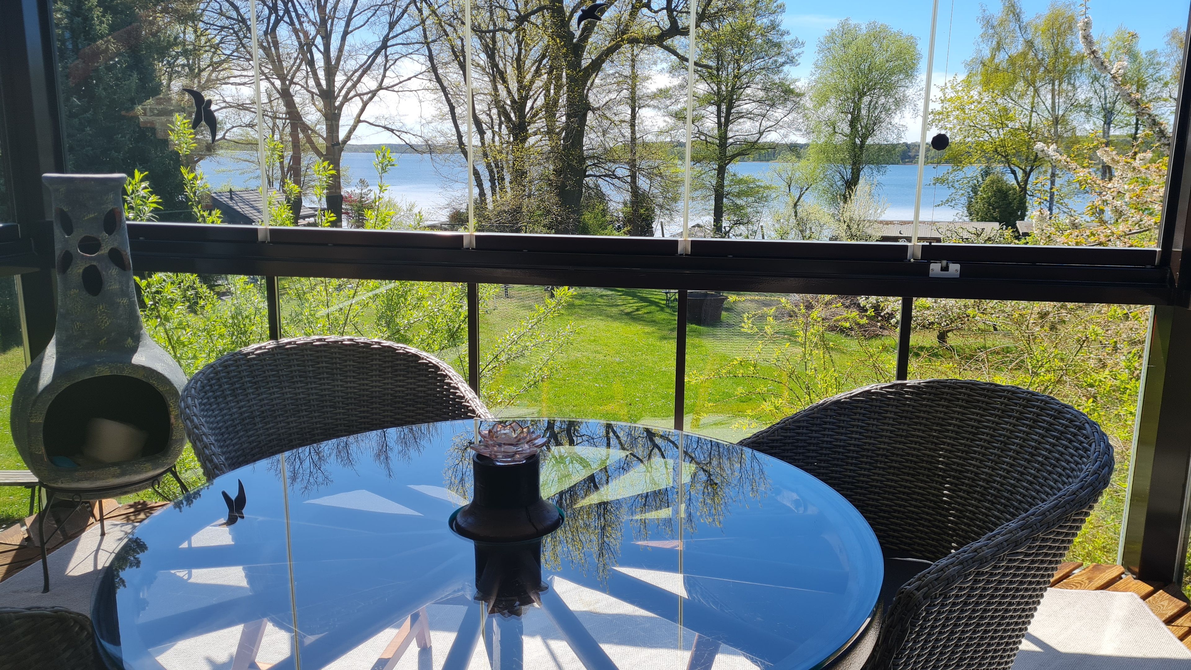 A balcony with wicker chairs and a glass table, overlooking a lake and greenery.