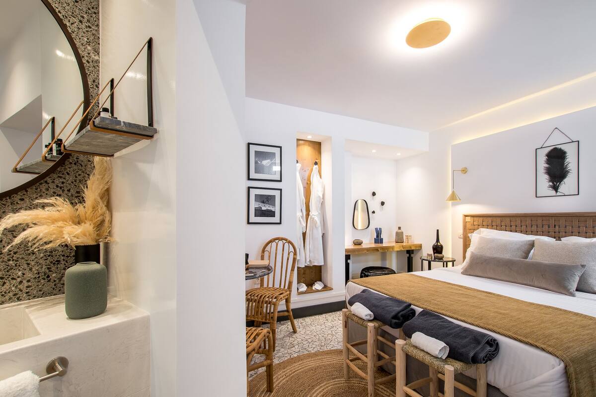 Una habitación de hotel moderna con una cama grande, un área de descanso con una silla y una mesa, un tocador con espejo y un baño con bañera y toallero.