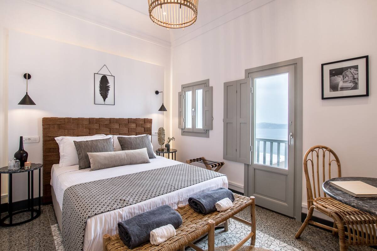 Una habitación de hotel con una cama grande, una mesita pequeña, un banco de mimbre y una puerta al balcón.