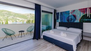 Una moderna camera d'albergo con un letto grande, due poltrone verdi, vista su un lago e montagne, e un quadro astratto dai colori vivaci appeso alla parete.