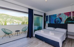 Una moderna camera d'albergo con un letto grande, due poltrone verdi, vista su un lago e montagne, e un quadro astratto dai colori vivaci appeso alla parete.