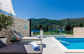 Un espace aménagé au bord de la piscine, avec des chaises longues, un mur en pierre et une vue sur un lac et des montagnes.