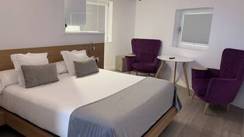 Ett modernt hotellrum med en stor säng, två lila fåtöljer, ett litet runt bord och ett fönster med persienner.