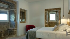A bedroom with a stone wall, a bed with white bedding, a red chair, and a small window. - Hotel Nande (As Neves)