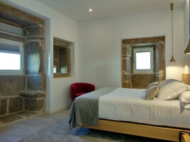 Un dormitorio con pared de piedra, una cama con ropa blanca, una silla roja y una ventana pequeña.