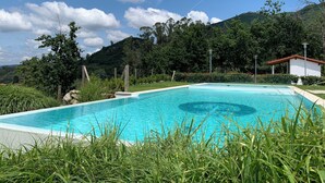 A rectangular swimming pool with clear blue water, surrounded by lush greenery and a mountain backdrop. - Hotel Nande (As Neves)
