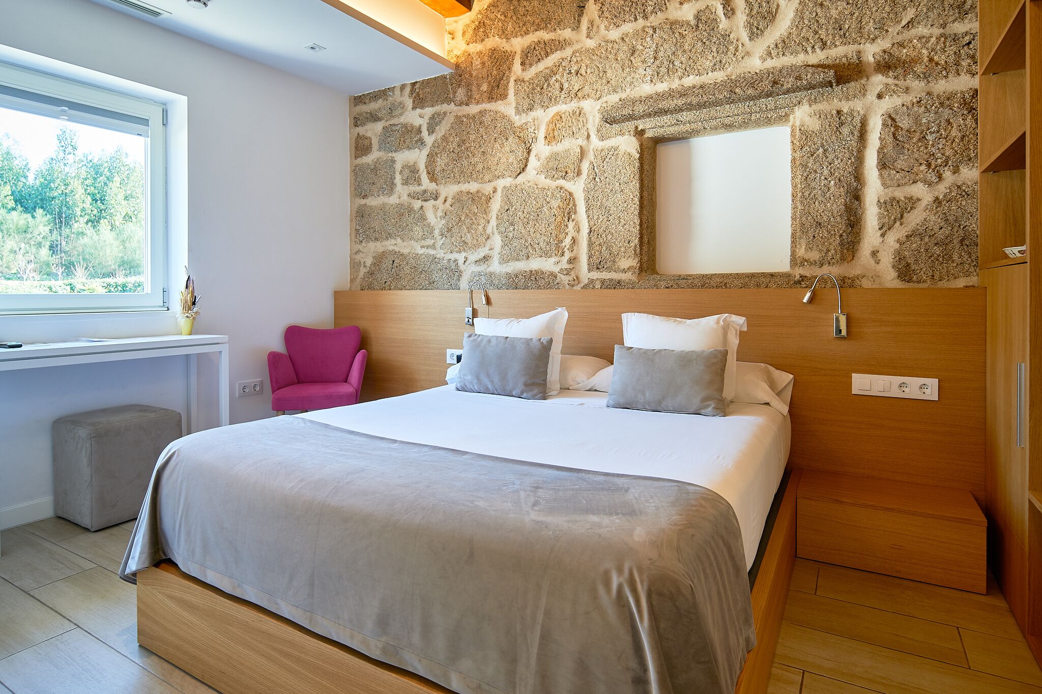 Un dormitorio moderno con pared de piedra, una cama grande, una silla rosa y una ventana con vistas a zonas verdes.