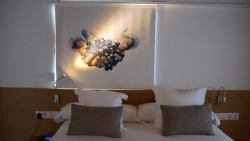 Habitación de hotel con una cama, dos almohadas, una mesita de noche con una lámpara y una obra de arte en la pared que representa uvas.