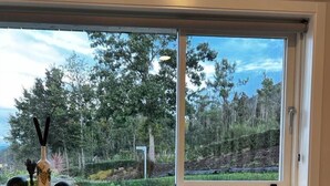 A dining table with a variety of food items and a bottle of juice, set against a window with a view of trees and hills. - Hotel Nande (As Neves)