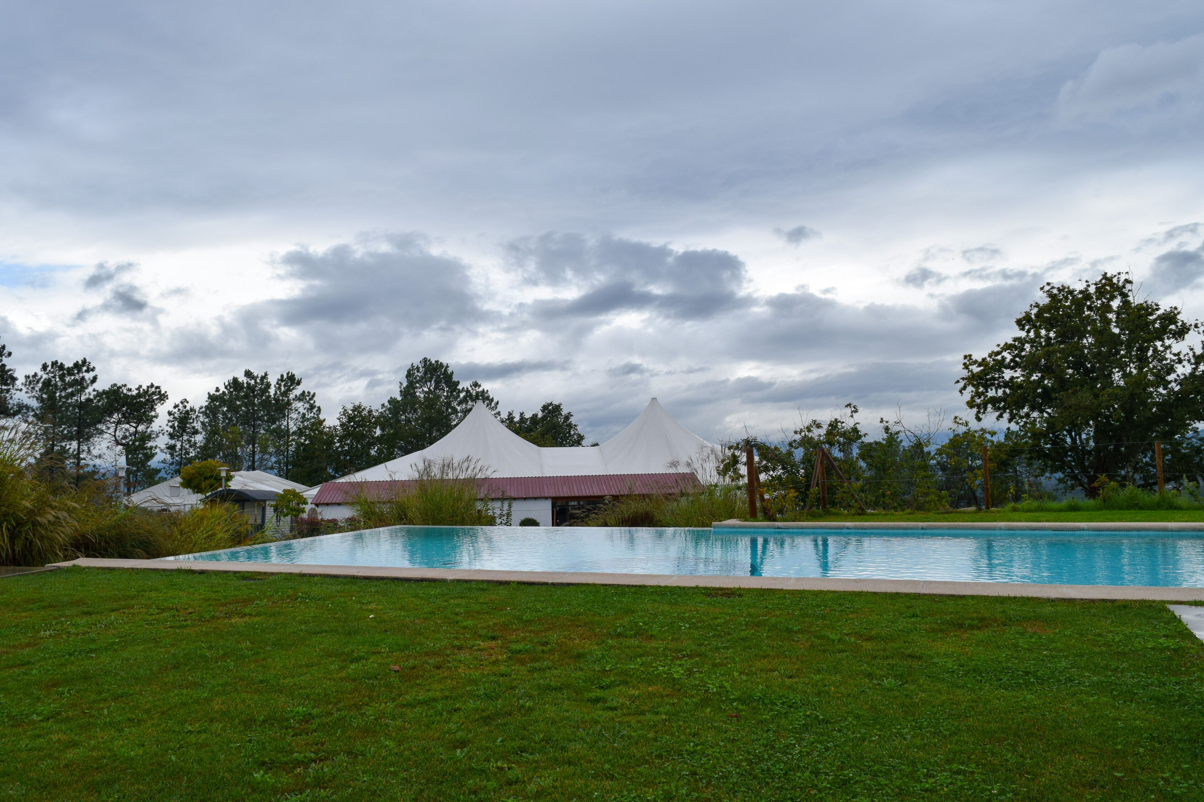 A swimming pool with a grassy area in front, surrounded by trees and a white tent-like structure.