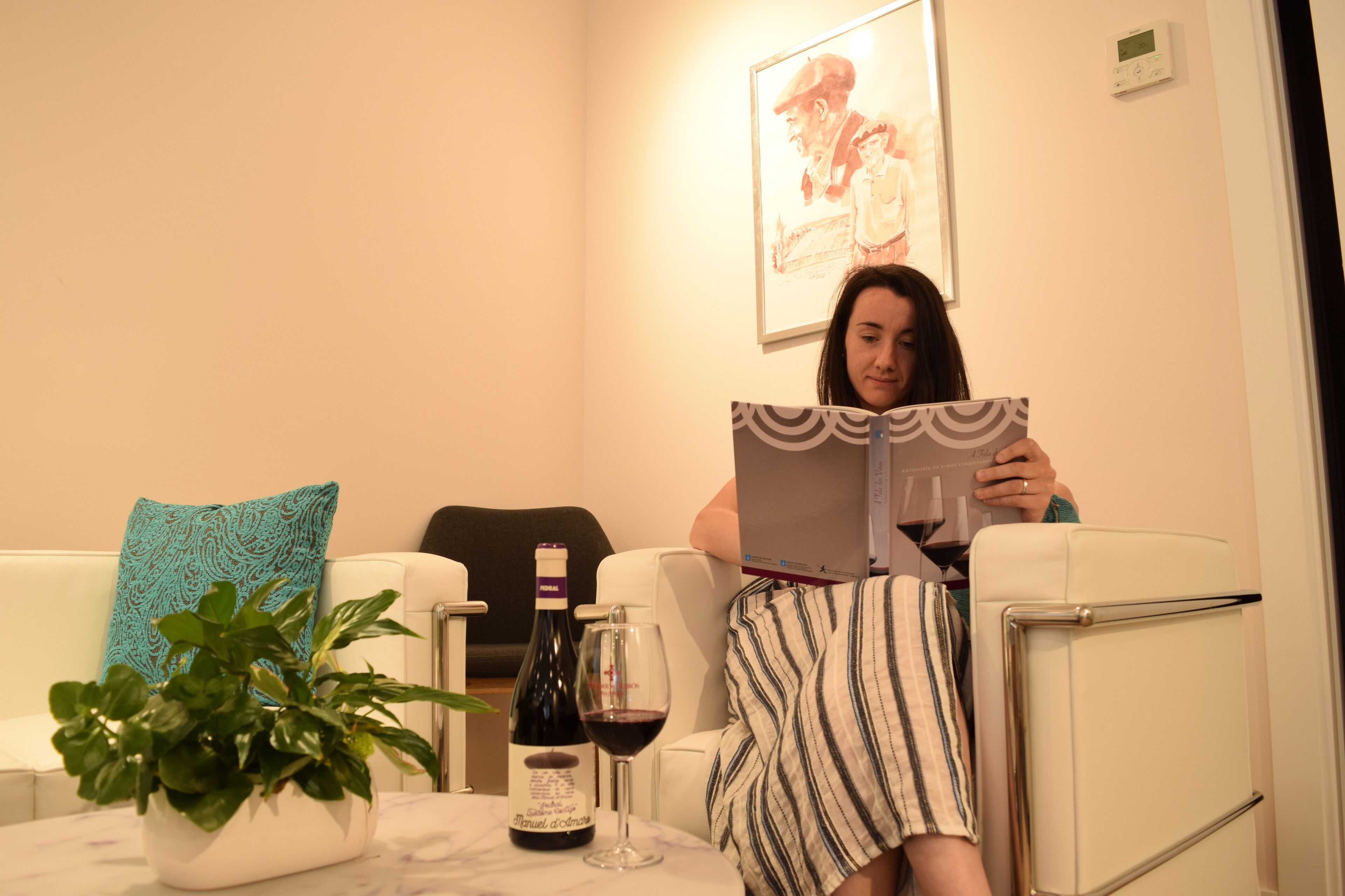 Uma pessoa sentada num sofá branco, com um livro, uma garrafa de vinho e um copo sobre uma mesa.