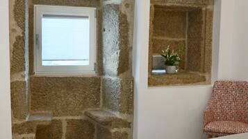 A bedroom with stone walls, a window, a bedside table with a plant, and a patterned chair.