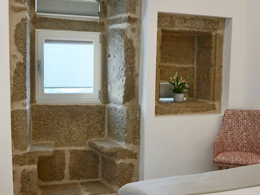 A bedroom with stone walls, a window, a bedside table with a plant, and a patterned chair.