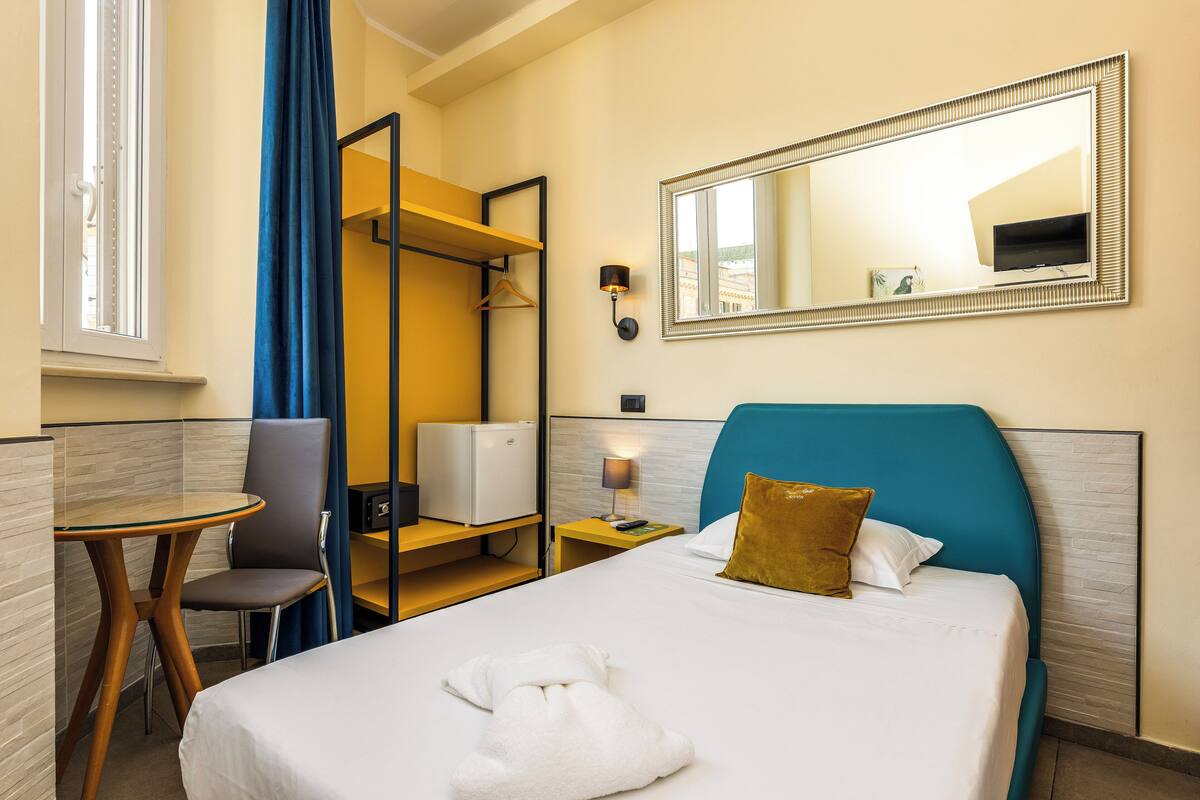 A hotel room with a bed, a desk, a chair, a mirror, and a wall-mounted TV.