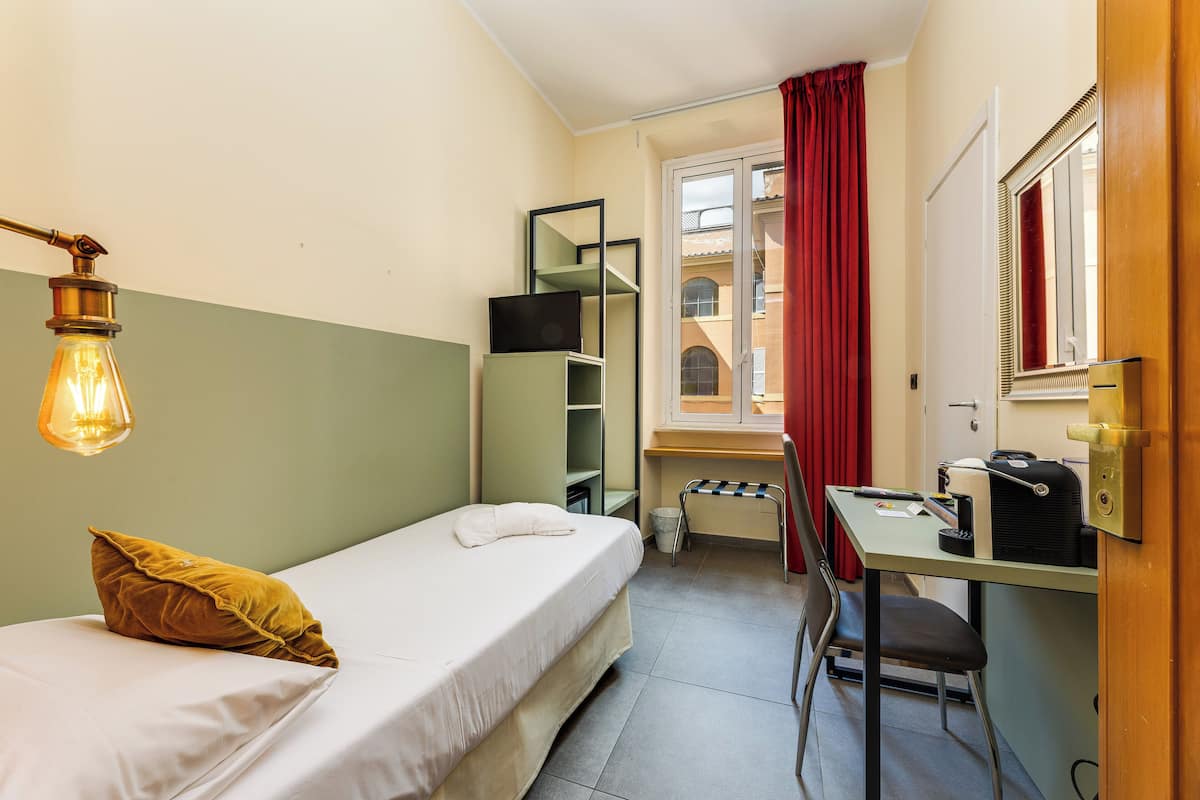 A small, compact room with a single bed, a desk with a chair, a mirror, and a window with red curtains.