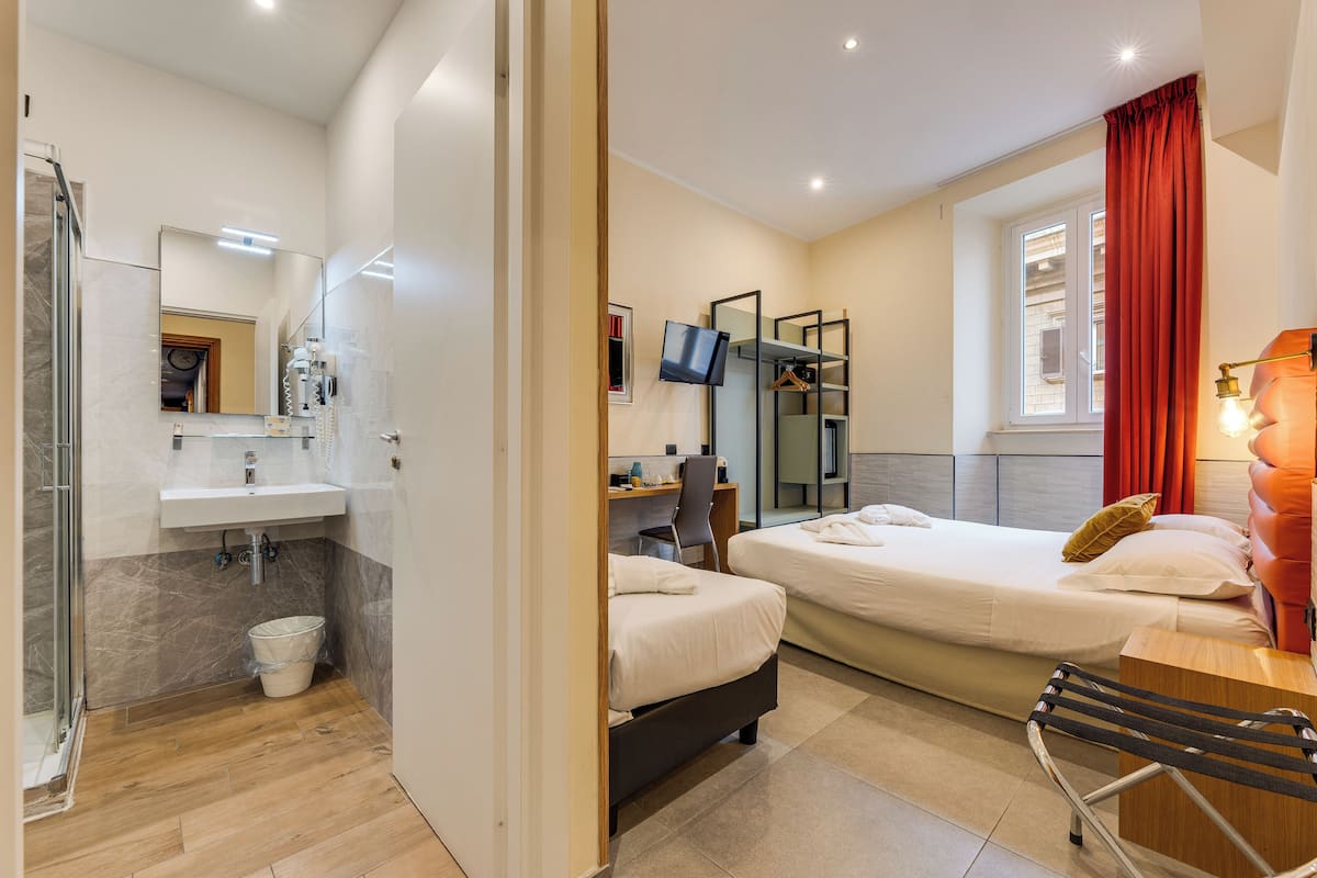 A modern hotel room with a shower, sink, and a bed with a desk and chair.
