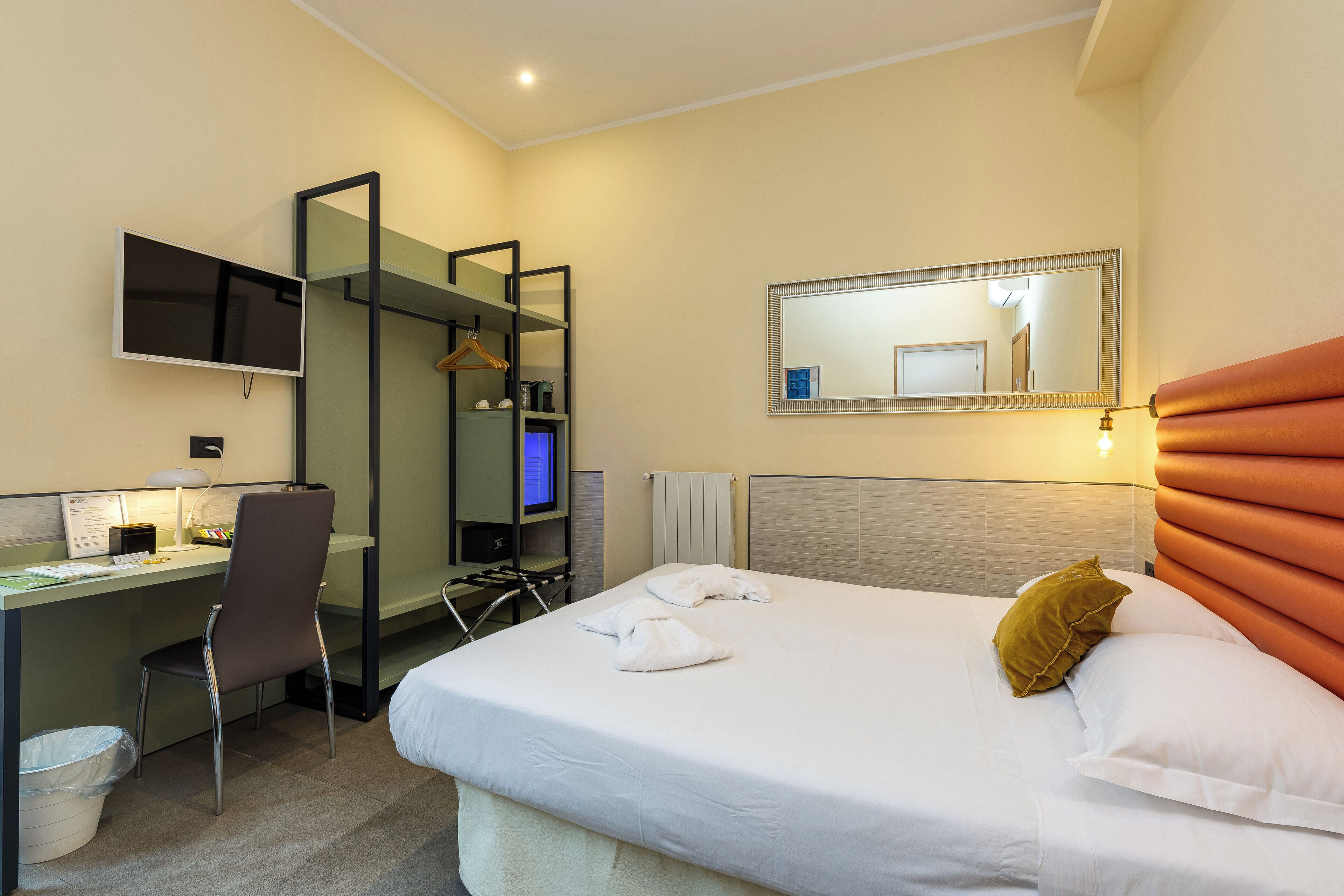 A hotel room with a bed, a desk with a chair, a TV, a mirror, and a radiator.