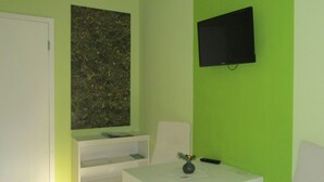 A room with a green wall, a flat-screen TV mounted on it, a white table with a vase, and a white chair. - Willischza (Burg (Spreewald))