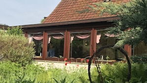 A house with a red-tiled roof, large windows with curtains, and a garden with various plants and flowers. - Willischza (Burg (Spreewald))