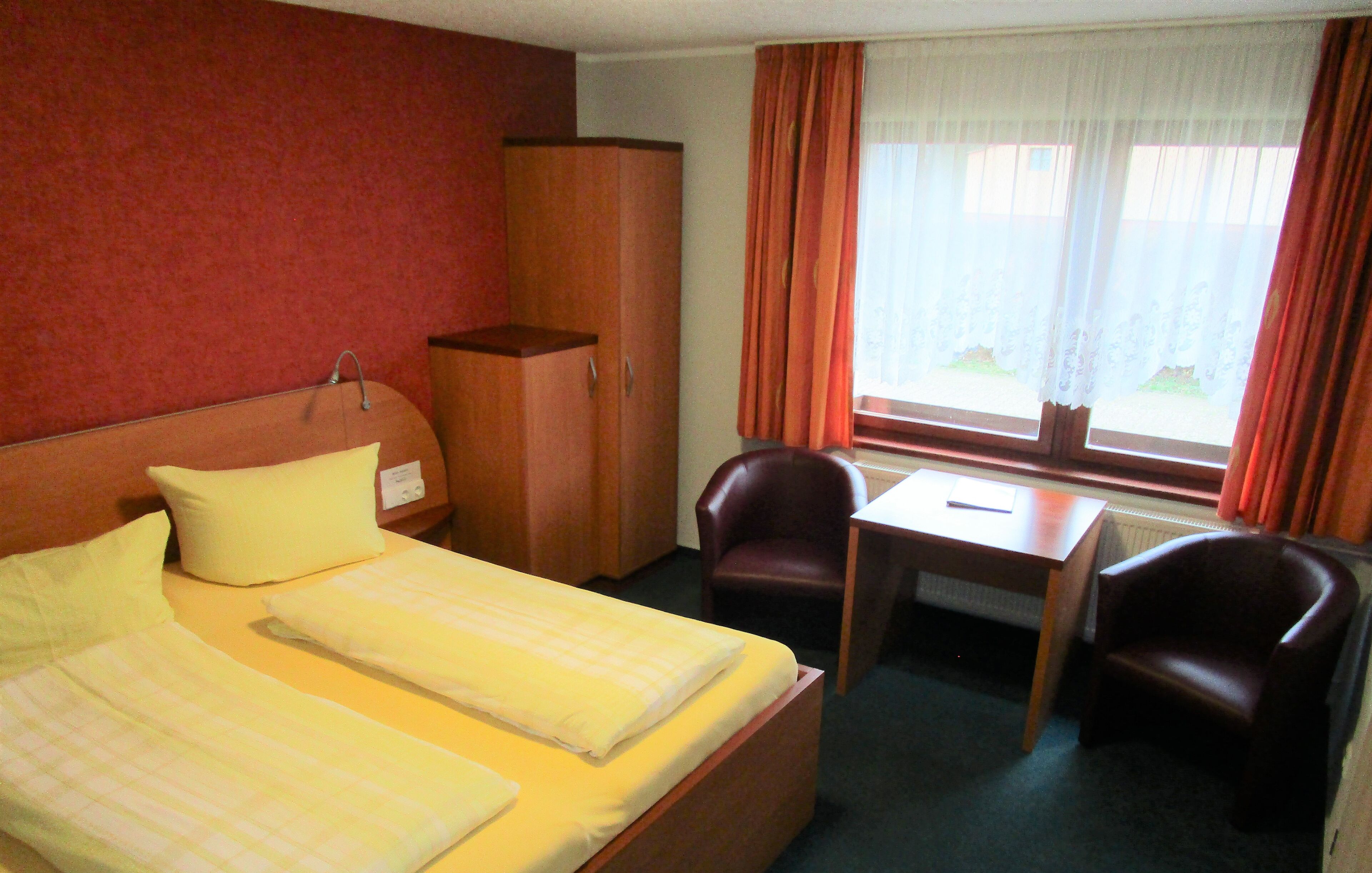 Ein Hotelzimmer mit zwei Betten, einem Schreibtisch, zwei Stühlen, einem Kleiderschrank und einem Fenster mit Vorhängen.