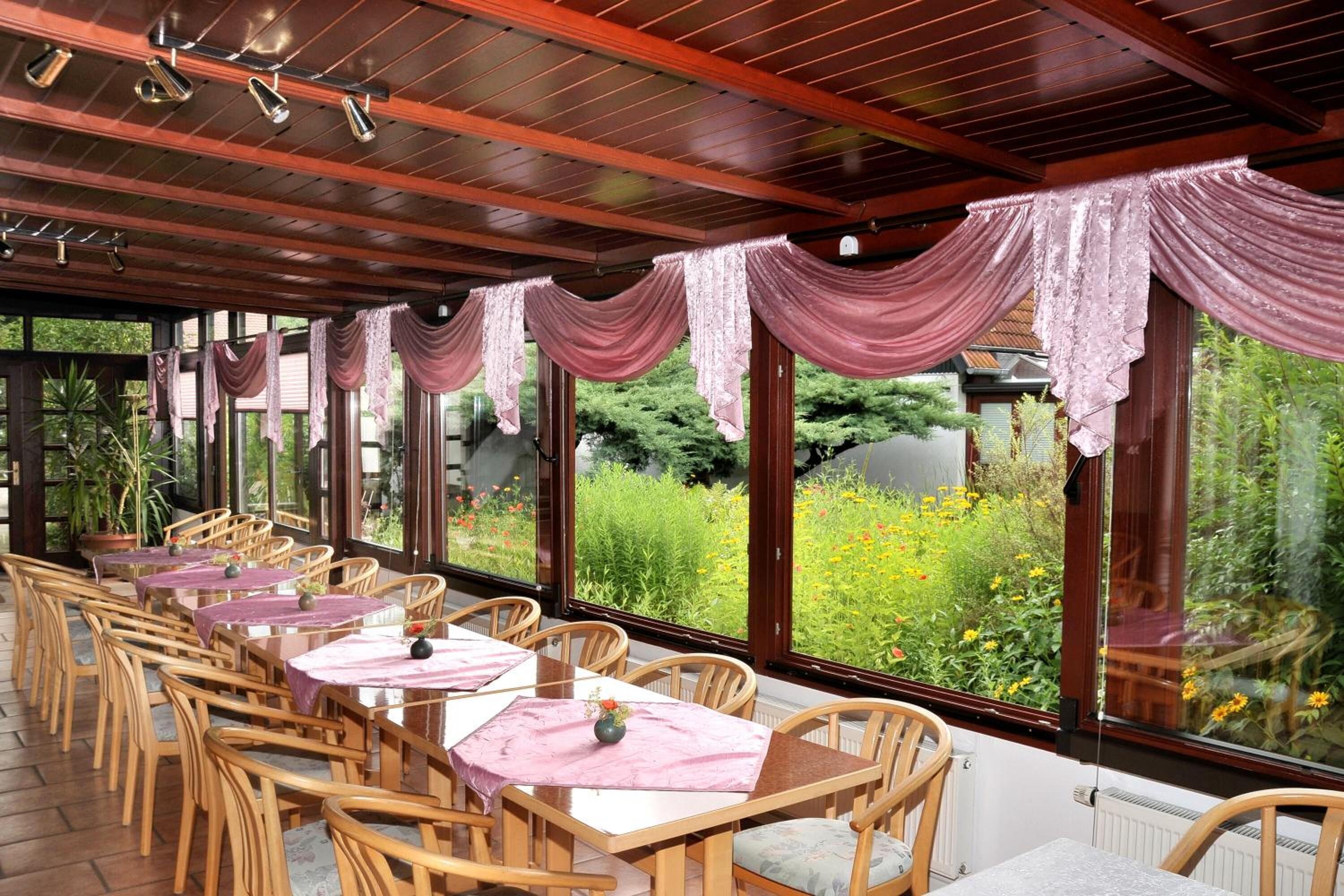 A covered dining area with wooden tables and chairs, large windows with curtains, and a view of a garden.