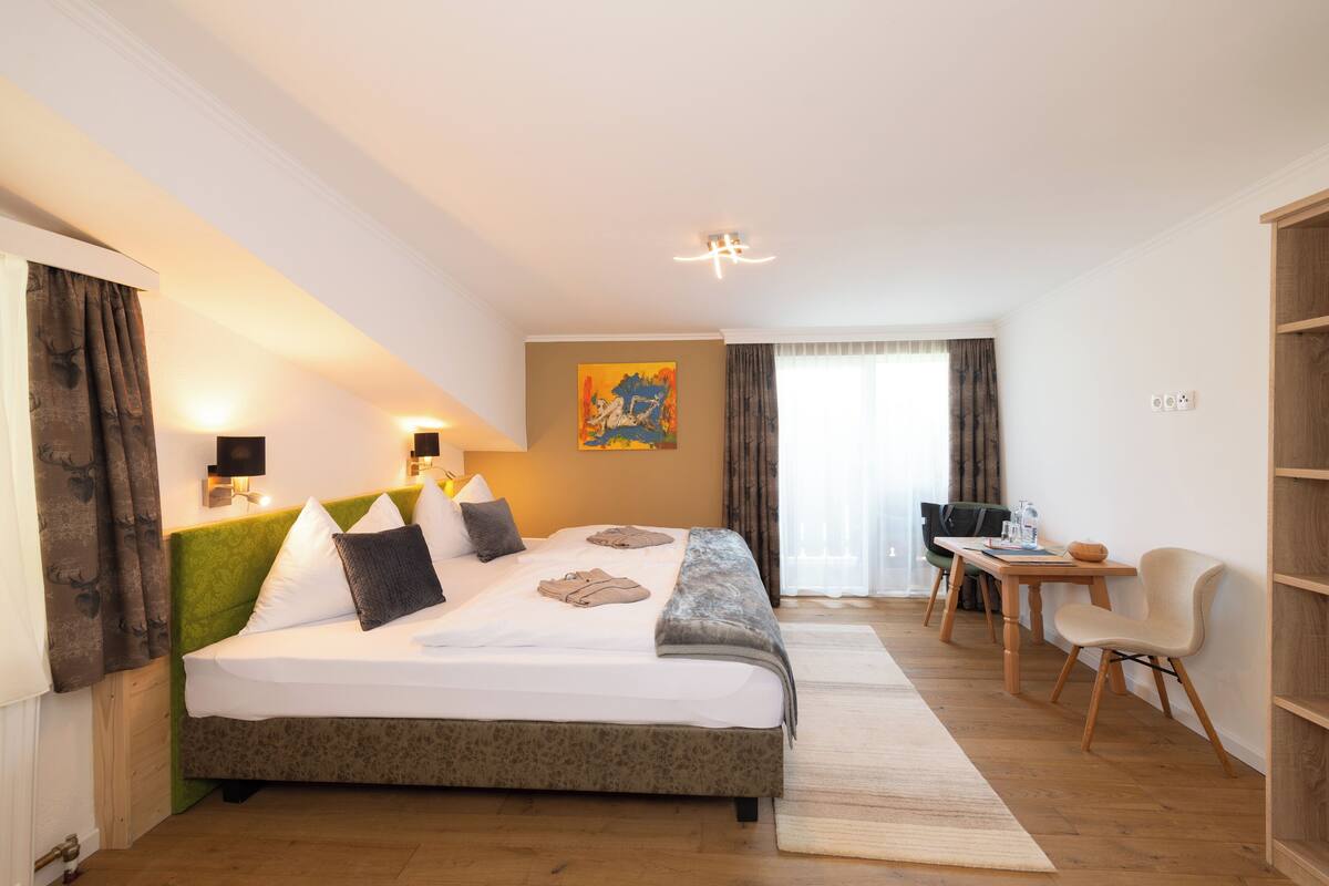 Een hotelkamer met een groot bed, een bureau en een stoel. Er hangt een schilderij aan de muur en er is een raam met gordijnen.